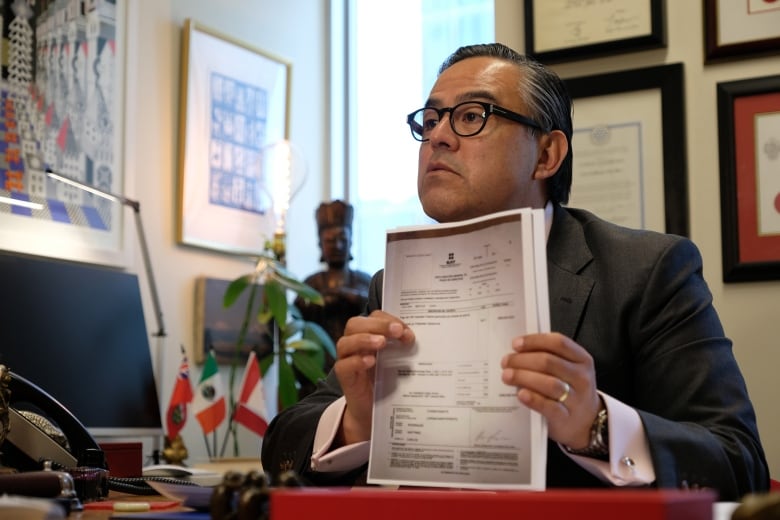 A man seated at a desk holds up a sheaf of photocopied paper. 