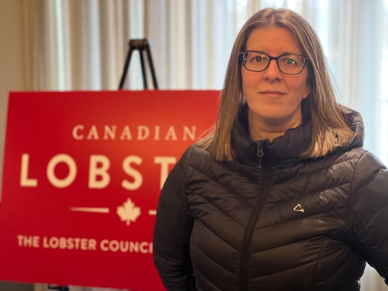 A woman with medium length hair and glasses wears a black jacket and stands in front of a lobster council of canada sign.