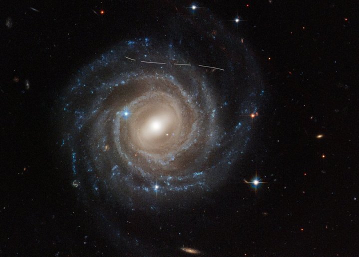 This NASA/ESA Hubble Space Telescope image of the barred spiral galaxy UGC 12158 looks like someone took a white marking pen to it. In reality it is a combination of time exposures of a foreground asteroid moving through Hubble’s field of view, photobombing the observation of the galaxy. Several exposures of the galaxy were taken, which is evidenced by the dashed pattern.