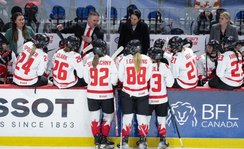 Four coaches stand behind the bench giving directions to a women's hockey team.