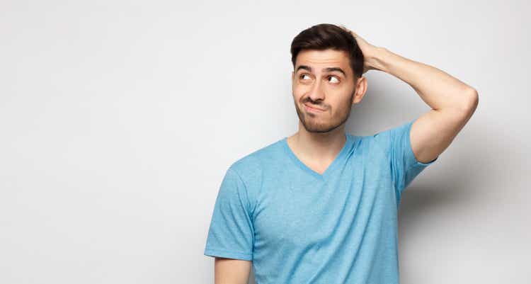 Young doubtful man thinking, scratching head and trying to find solution, isolated on gray background