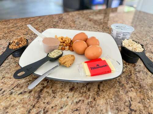 a spread of food containing granola, protein powder, hemp seeds, peanut butter, cheese, nuts, eggs, oats, and yogurt depicting 100 grams of protein