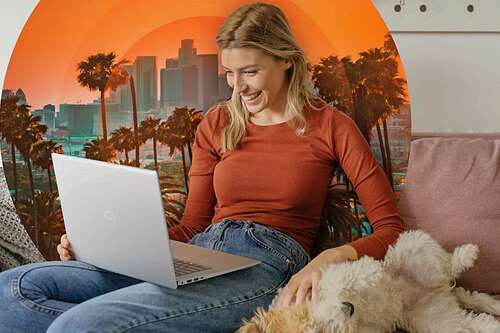 A woman sitting on the couch using an Inspiron laptop while petting her dog.