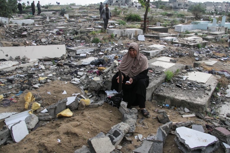 A woman in a headscarf sits in a field with stone graves and concrete pieces of debris.