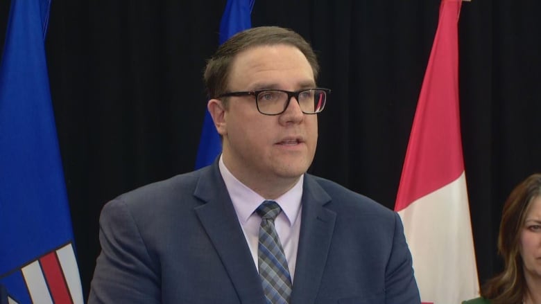 A man with short brown hair, glasses and a suit stands before a microphone.