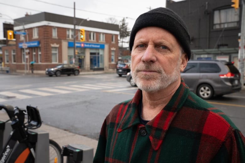 A man in a toque and plaid jacket stands in front of a bank.