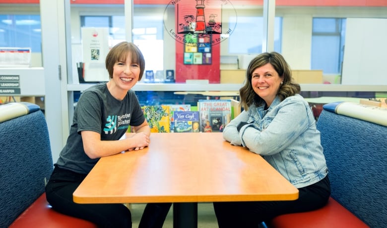 Two smiling woman sit on banquettes across from each other with their forearms resting on a table. 