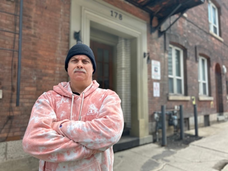 Someone in a tie-dye hoodie crosses their arms in front of a low-rise brick building.