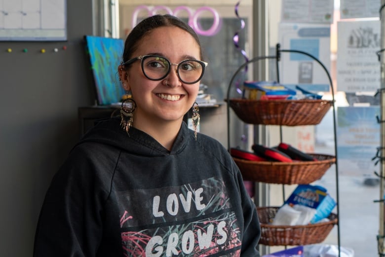 A person wearing glasses and a hoodie that says "Love Grows" stands inside, smiling. Behind them are baskets filled with sanitary supplies and naloxone kits.