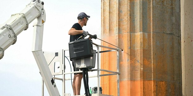 Ein Mann steht auf einer Hebebühne und reinigt mit einem Hochdruckreiniger eine Säule des Brandenburger Tors, die mit orranger Farbe besprüht wurde