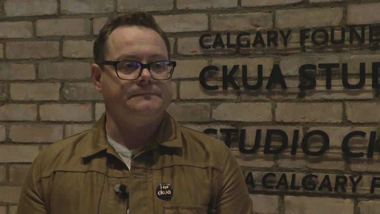 A white man with short dark hair and large glasses is standing indoors, in front of a brick wall. He is wearing a casual jacket with a 'CKUA' button on his left breast pocket.