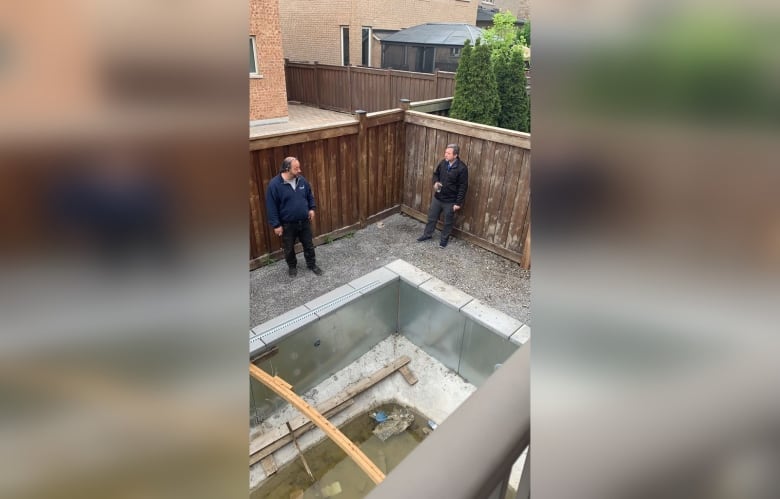 Two men are speaking in the backyard of a home next to an empty, unfinished pool.