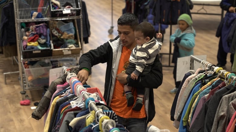 A man looks through clothing racks while holding a baby. They're in a church basement.