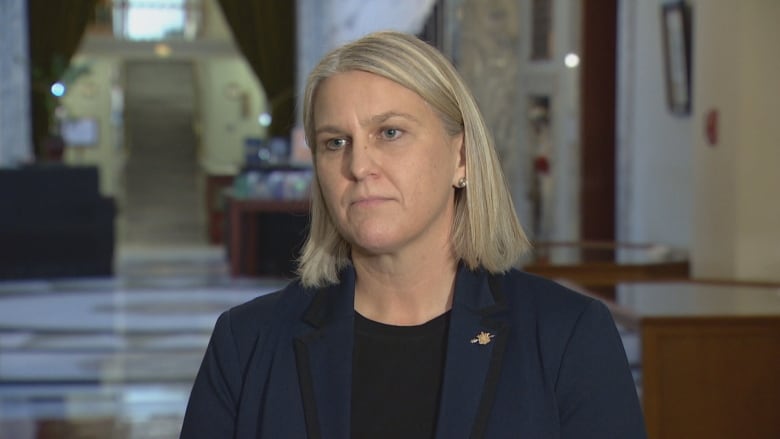 A woman with straight bright blonde hair in a bob, looks off to the left of the frame. She's wearing a black shirt and a blue blazer.