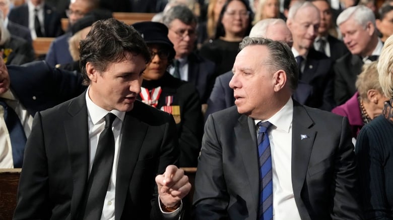 Two men interact with each other. One is wearing a black suit, the other is wearing a gray suit.