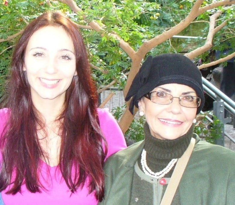 A young woman with long auburn hair smiles while standing next to an older woman wearing a black hat.