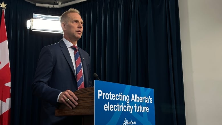 A blonde man stands at a podium with a blue sign that says "Protecting Alberta's electricity future."