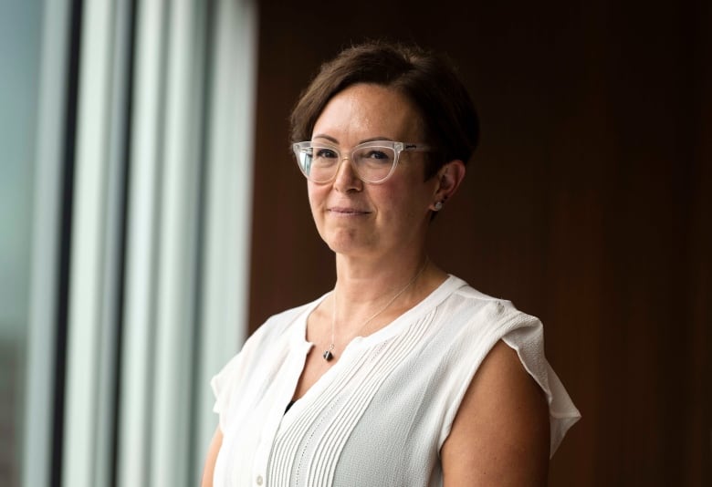 A woman in a white shirt and glasses stands before a window.