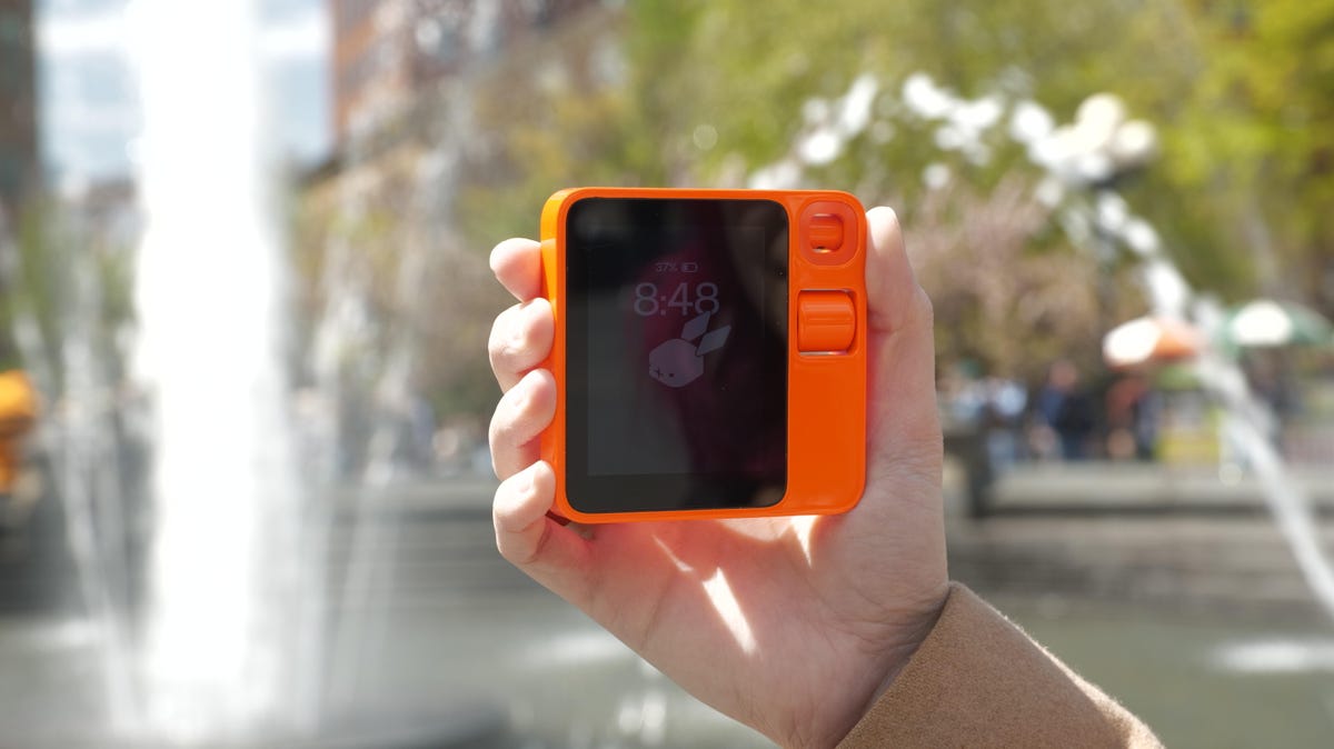 The Rabbit R1 being held up in front of a fountain.