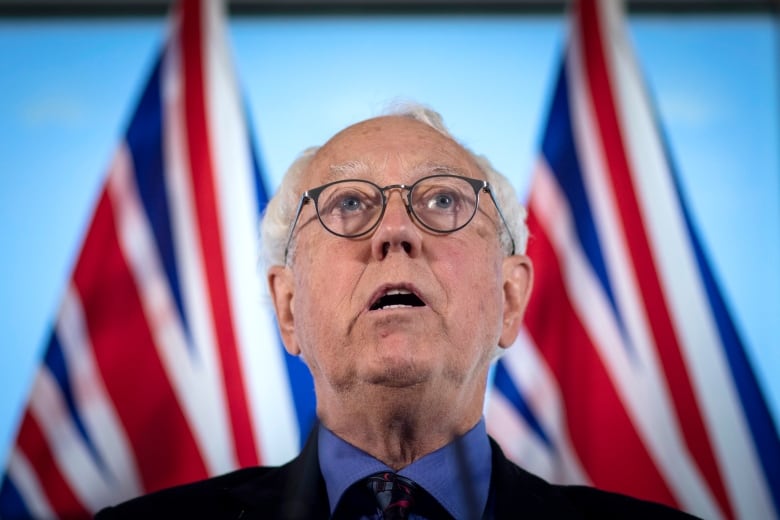 A white man with round glasses speaks in front of B.C. flags.