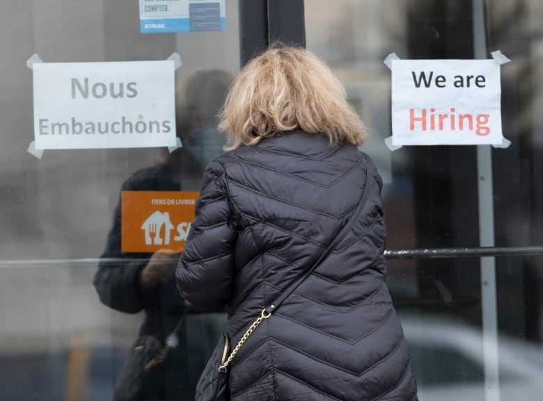 A woman is seen from behind going through a door that has signs in both English and French that say the establishment is hiring.