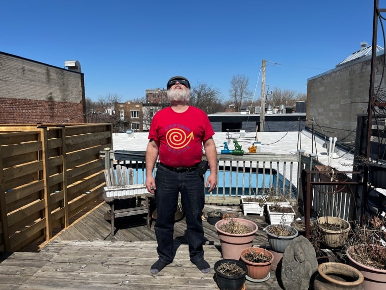 A man in a red shirt and eclipse glasses peers at the sky