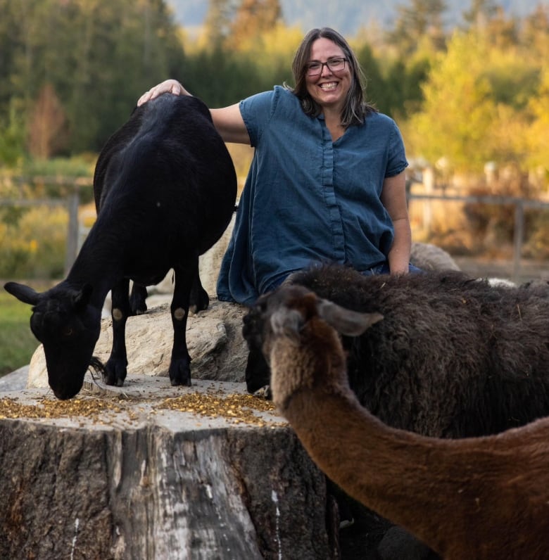 A woman leans on a goat while smiling the camera. Also in the frame is an alpaca.