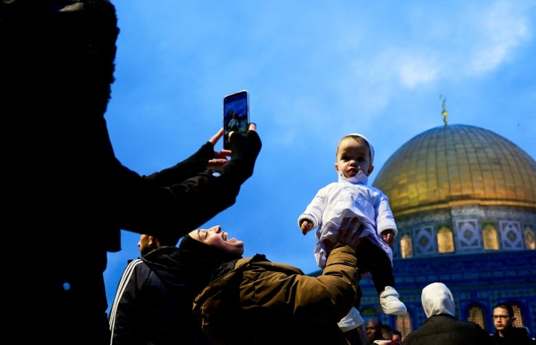 A woman holds up a small child as another takes a photograph of the child, with a domed religious structure shown in the background. 