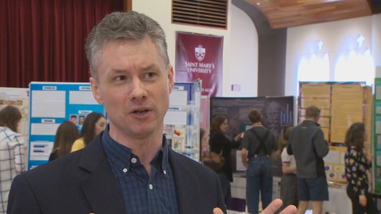 A man in a black jacket and dark shirt speaks to the camera at a science fair.