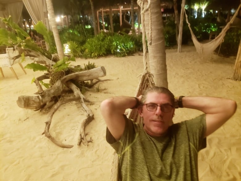 A man on a beach leans back against a tree. 