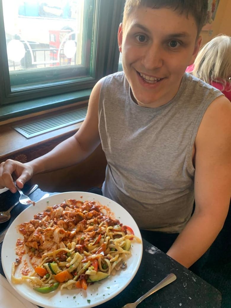 A boy in a grey shirt sits at a table with a plate of pasta in front of him. 