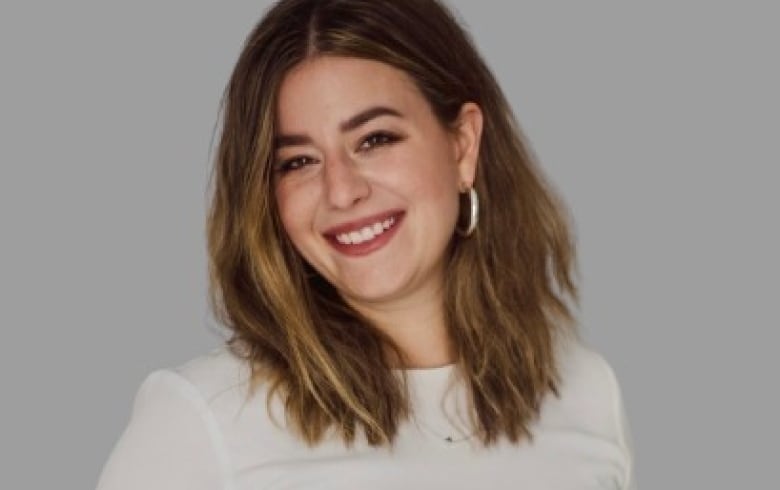 A woman stands and poses for a photo. she us wear a white top and dark pinstripe pants. She has her hands in her pockets and is smiling for the camera. 