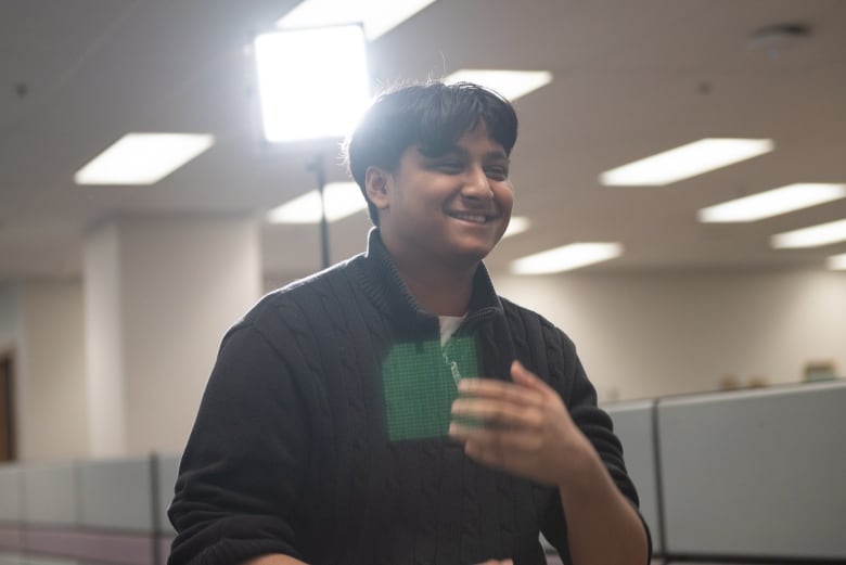 A teen boy gestures and smiles broadly to someone off camera to the right, with bright television lights in an indoor space seen behind him.