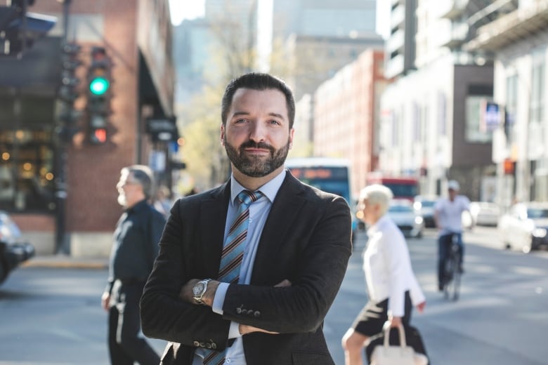 A man with a beard and moustache, wearing a suit and tie, stands with his arms crossed on a city sidewalk, with busy streets, buildings and pedestrians seen in the background.