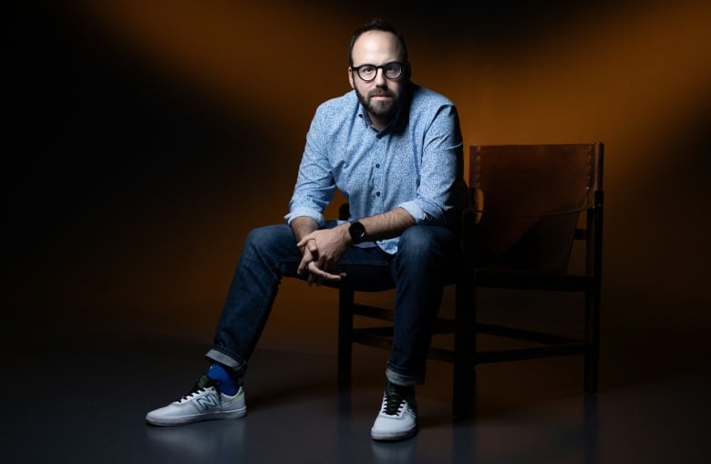 A bearded man with glasses wearing jeans, sneakers and a blue button-up shirt sits on a chair and poses for a photo. 