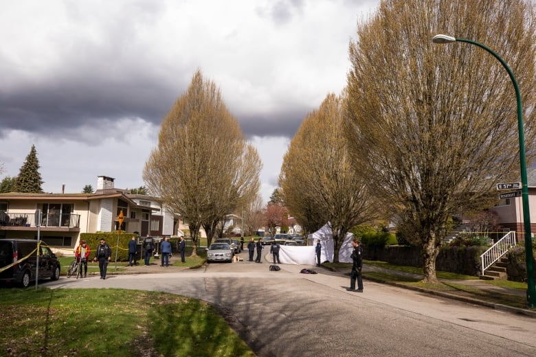 A wideshot image of a residential street with almost a dozen police investigators and a white tent.