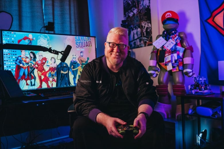 Photo of a man wearing a black jacket and holding a video game controller. A large computer screen and mannequin wearing a Mario hat is in the background.