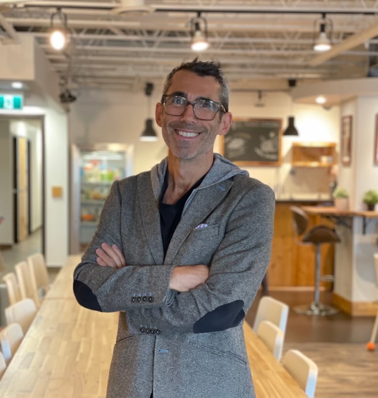 A man wearing a grey sport coat over a hoodie smiles as he poses for a photo in front of a long table in a space that has exposed metal rafters and a kitchen in the background.