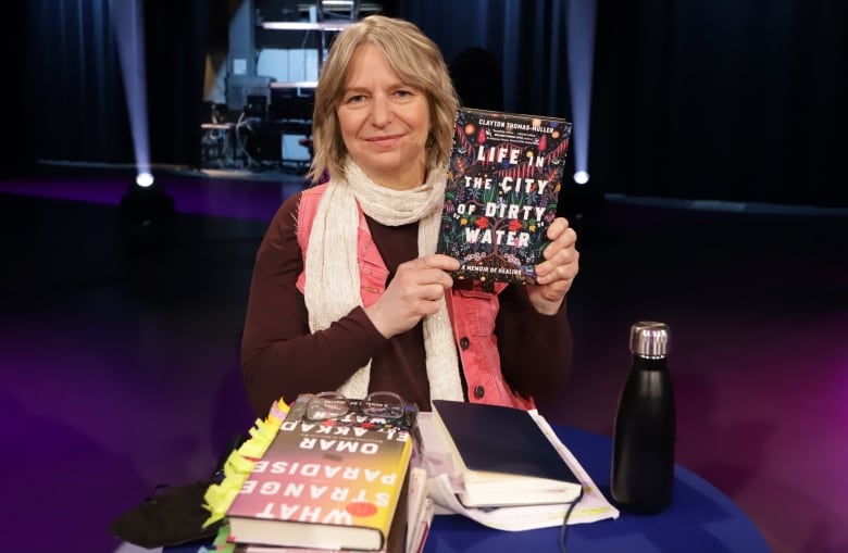 A blonde white woman holds up a book called Life in the city of dirty water.