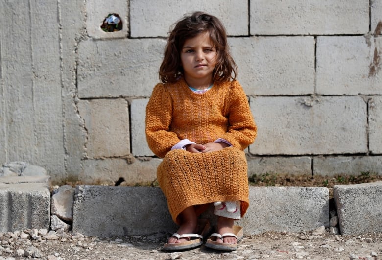 A small girl in an orange dress sits in front of a plain brick wall.