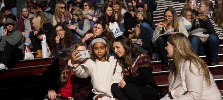 A trio of teen girls look at a smartphone while sitting outdoors on a steps. Other teens are seen sitting on steps in the background.