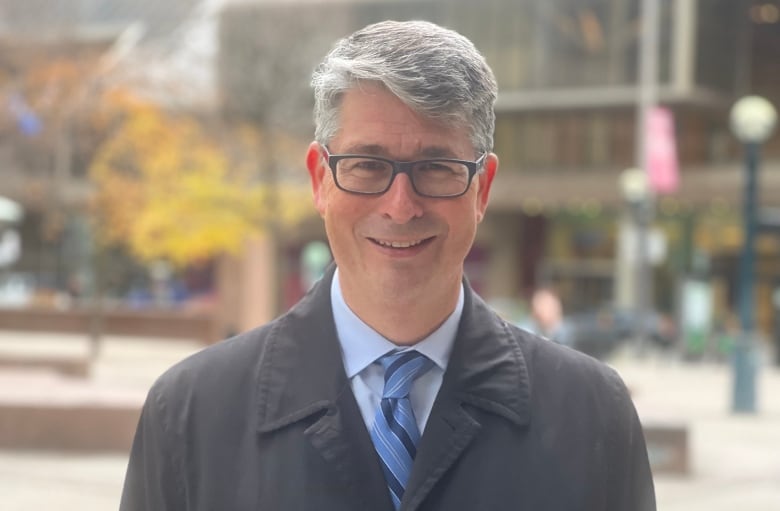 A photo of man with glasses grey hair with a black jacket, blue shirt and tie.