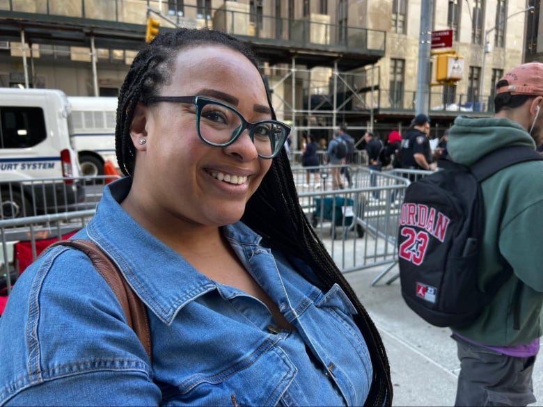 A woman wearing glasses and a blue shirt smiles while standing on the street.