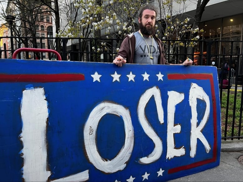 A bearded man standing behind a large sign that reads loser