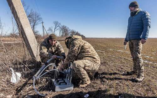 Two soldiers work on a piece of machinery consisting of a metal rectangular square with three heavy attached cables, as well as three vertical pieces coming out of it, while another man looks on.