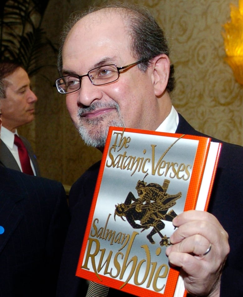 A man with glasses and a bald spot on top of his head and a white, short beard, holds a book up in his left hand. The book has an orange border and the title "The Satanic Verses Salman Rushdie." 