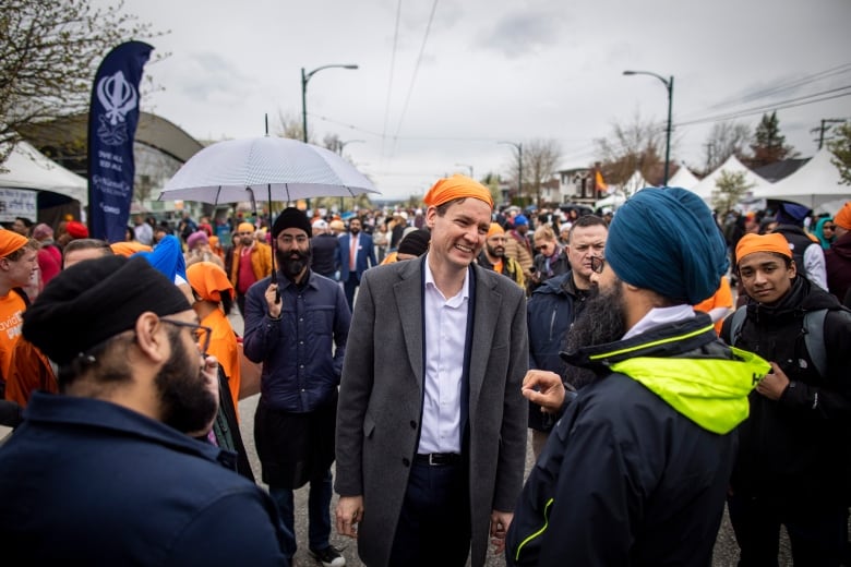 A white man wearing an orange head covering talks to a Sikh man with thousands of people behind him.