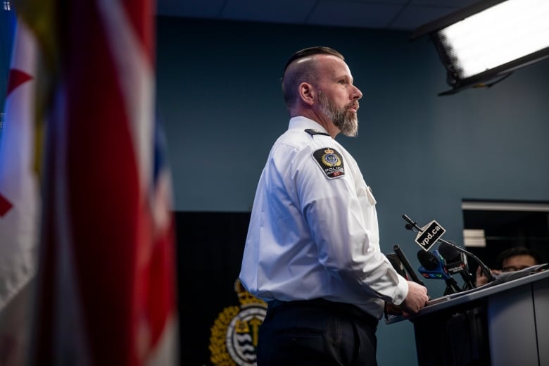 A police officer with a grey beard stands at a podium as he holds a news conference. 