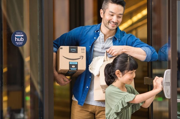 A parent holding an Amazon package under their right arm while their child holds a door open for them.