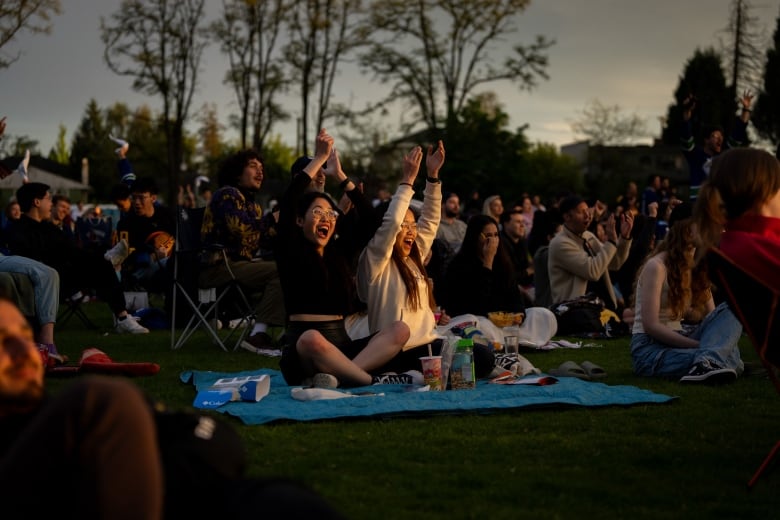 A crowd of people sit on blankets on the grass at dusk, clapping and smiling.